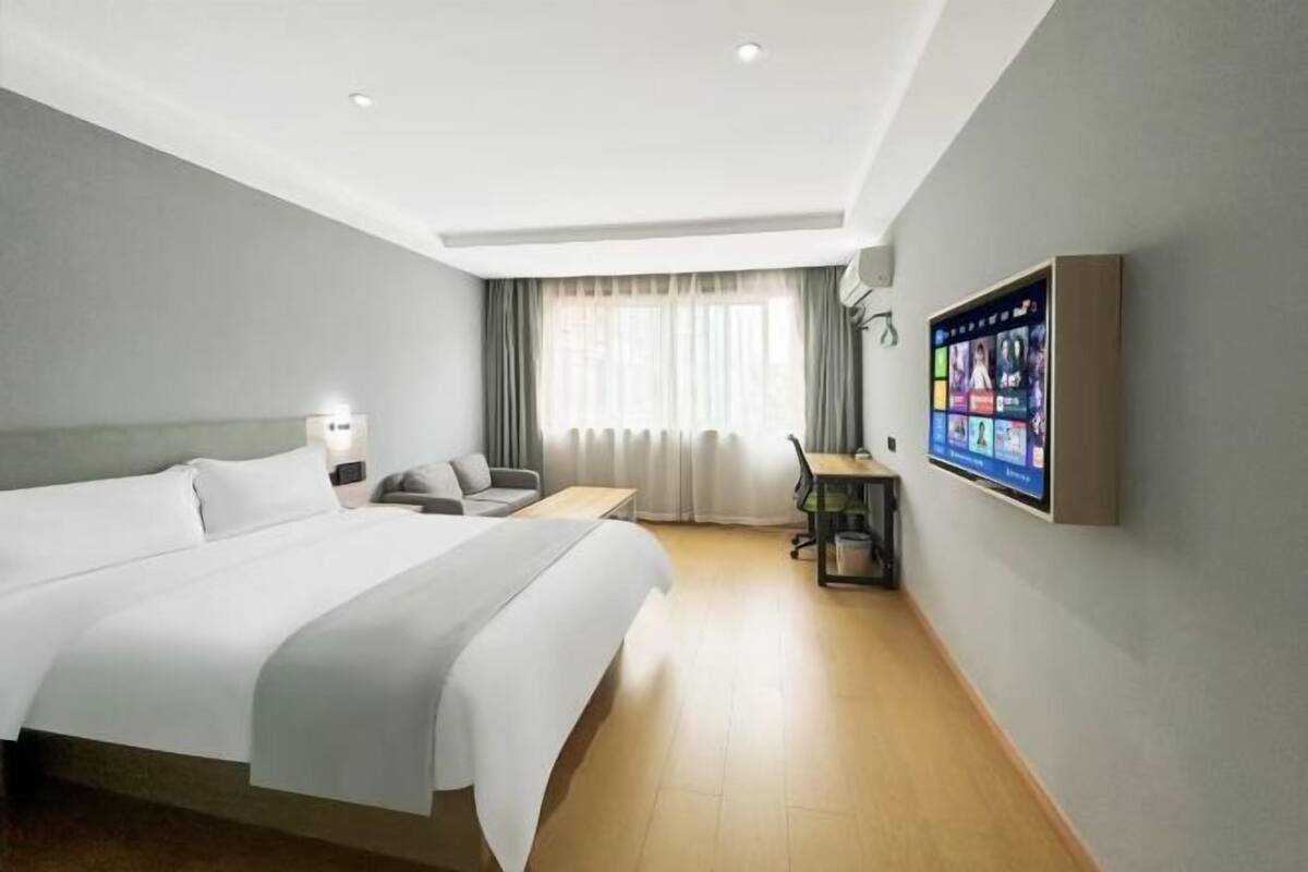 Habitación de hotel con una cama grande, un escritorio, una silla, un televisor montado en la pared y una ventana con cortinas.