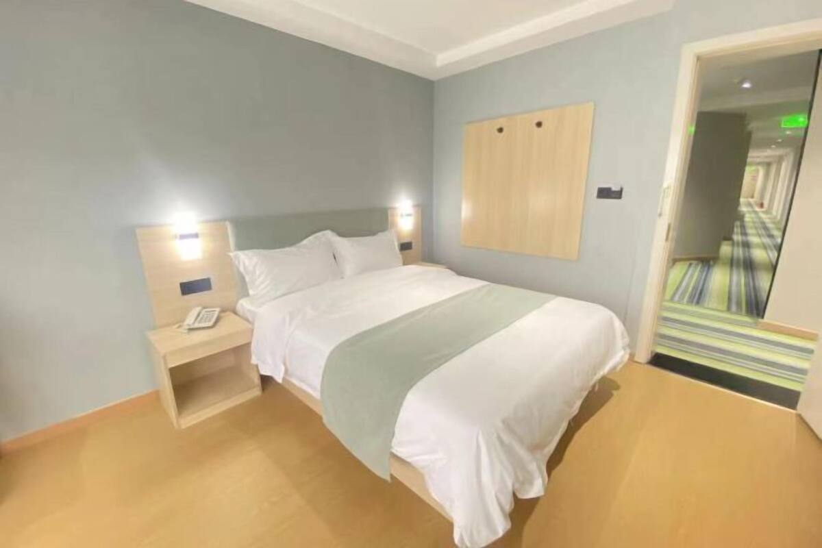 Habitación de hotel con cama, mesita de noche con teléfono, lámpara de pared y puerta que da a un pasillo.