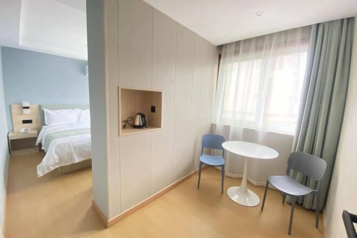 Una habitación de hotel con una cama, una mesita pequeña con dos sillas, una mesita de noche con una lámpara y una ventana con cortinas.