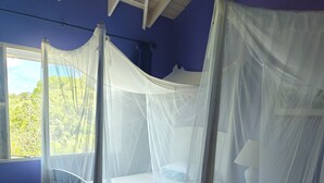 Una habitación con dos camas cubiertas por mosquiteros blancos, suelo de madera y ventana con vista a la vegetación.