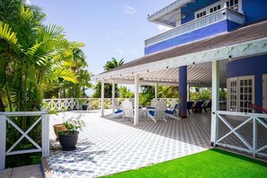 A covered outdoor seating area with chairs and tables, surrounded by a white fence and lush greenery. - Luxury Tobago Estate - Pool, Sports Court, Sleeps 15 (Patience Hill)