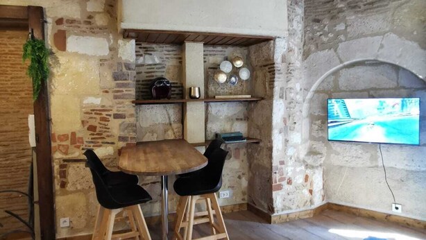 A rustic room with exposed stone walls, a wooden table with two chairs, a television mounted on the wall, and a blue sofa. - 
Chic Studio in Old Bergerac (Bergerac)