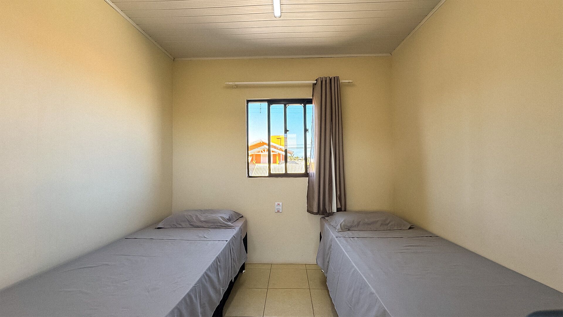 A small, single-room accommodation with two single beds, a window with curtains, and a view of buildings outside.