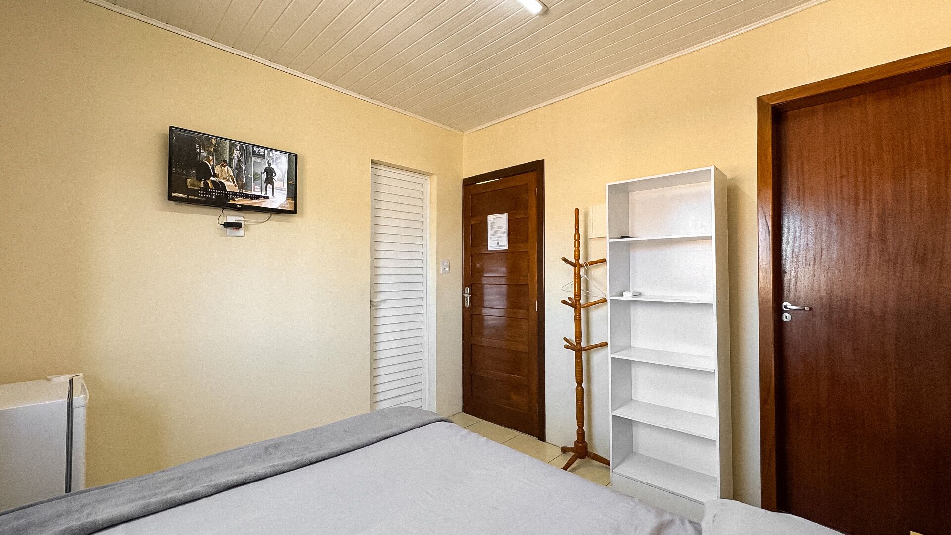 Un dormitorio con cama, televisión montada en la pared, una puerta de madera y una estantería blanca.
