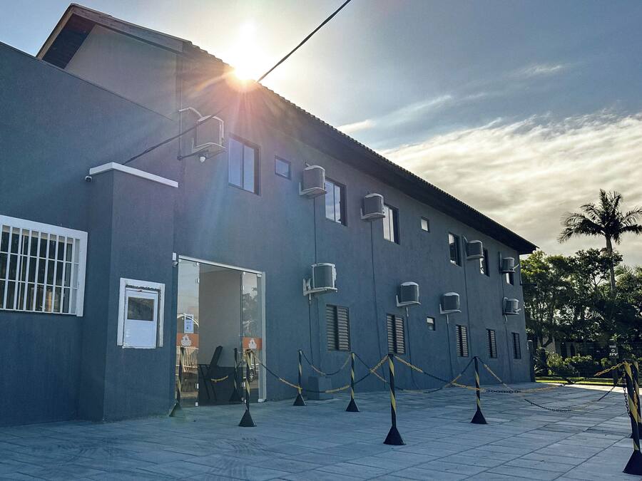 Un edificio moderno con techo plano, ventanas amplias y unidades de aire acondicionado. Hay cadenas de seguridad y conos frente a la entrada.