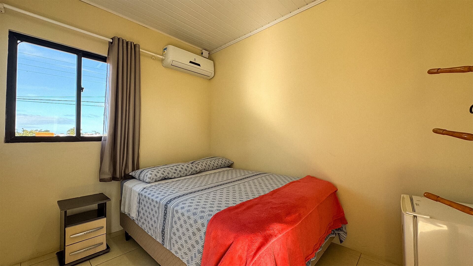 A bedroom with a bed, a window with curtains, an air conditioning unit, a small cabinet, and a radiator.