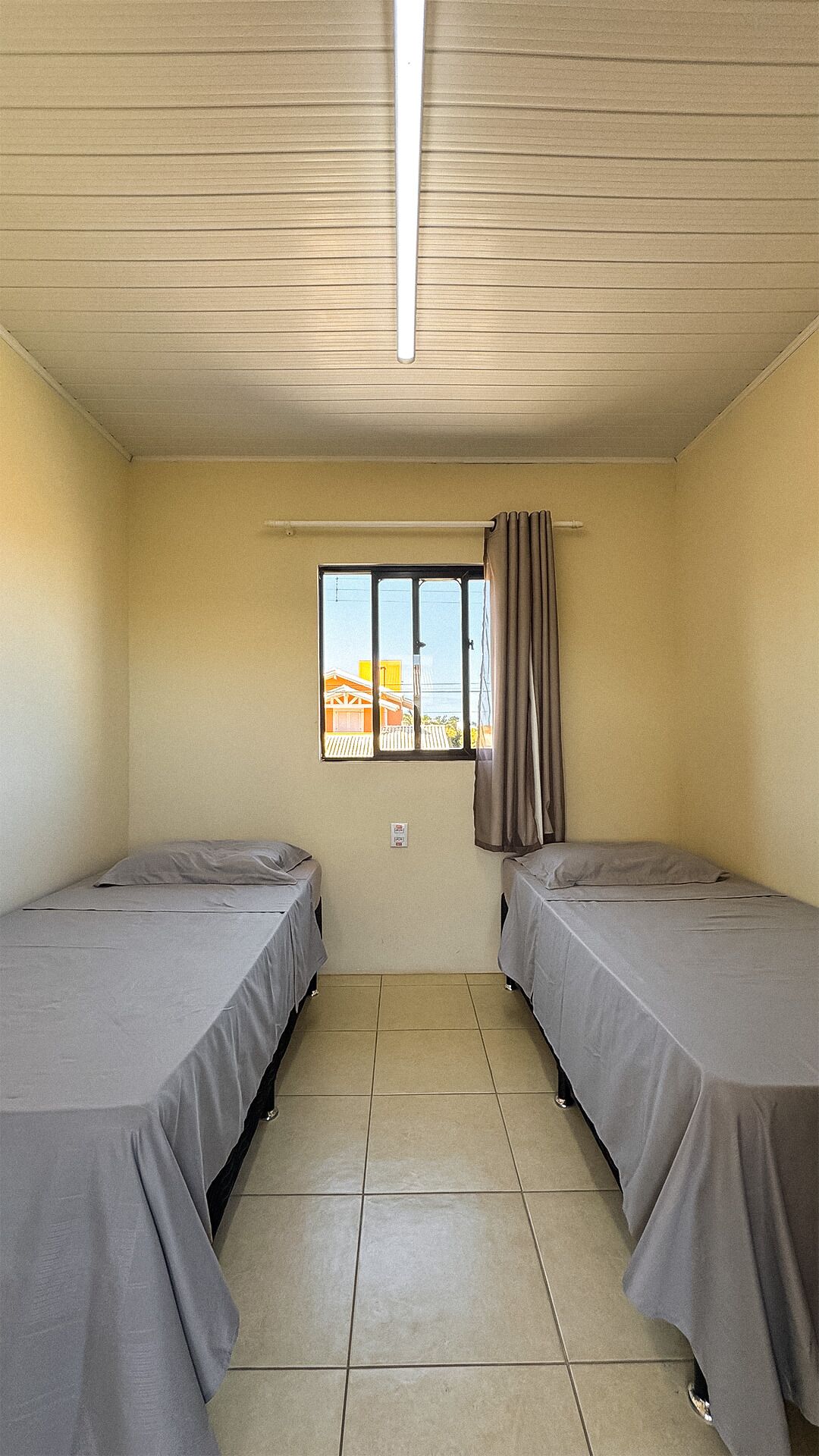 A small, single-room hotel room with two beds, a window with curtains, and a tiled floor.