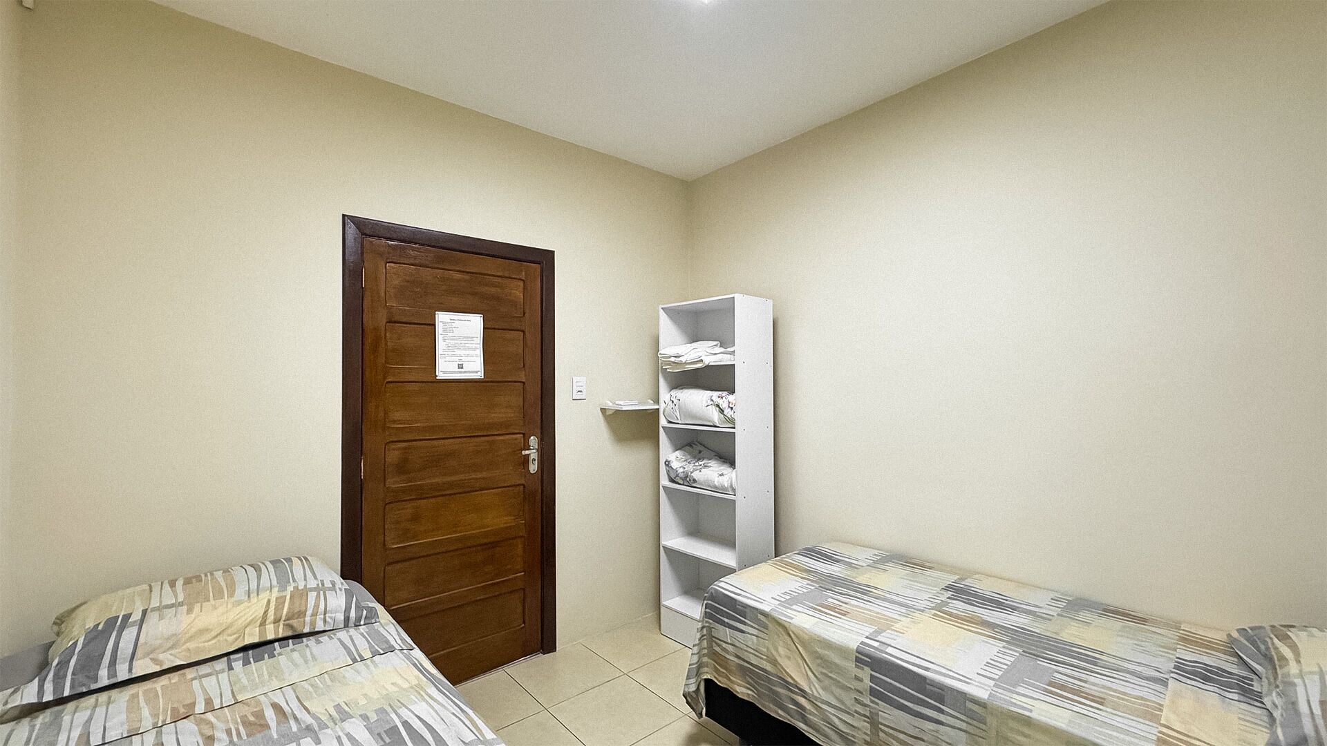 Una habitación pequeña y compacta con dos camas individuales, una puerta de madera y un estante blanco en la pared.