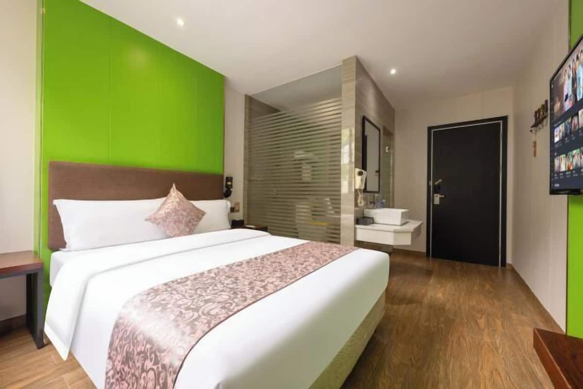 Una habitación de hotel moderna con una cama grande, una mesita de noche, un baño con lavamanos y espejo, y una pared verde decorativa.