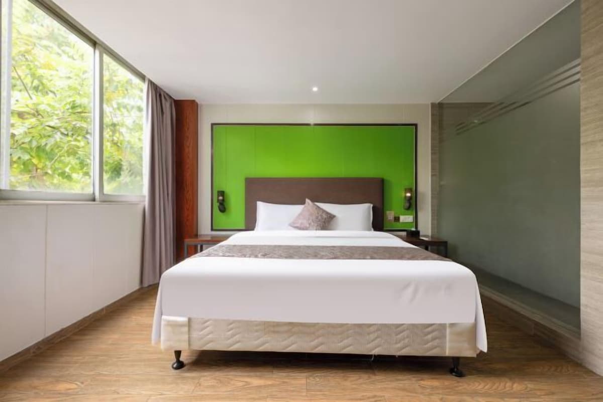 A modern hotel room with a large bed, green accent wall, and a view of trees through the window.