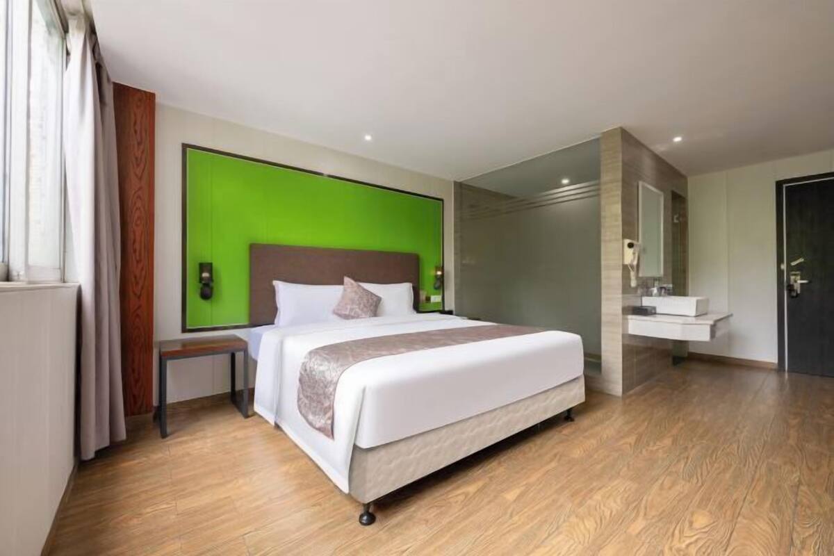 Una habitación de hotel moderna con cama, mesita de noche, pared de acento verde y un baño con espejo y lavamanos.