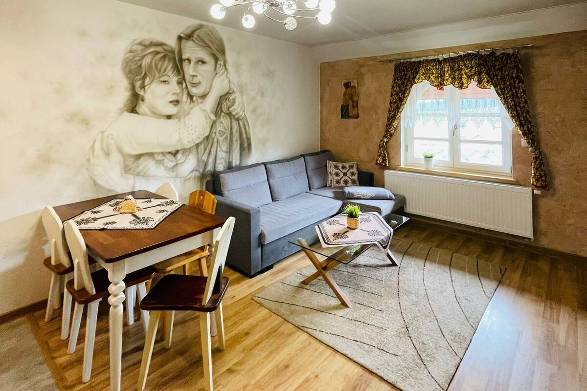 A living room with a sofa, a dining table with chairs, a chandelier, a window with curtains, and a wall with a mural.