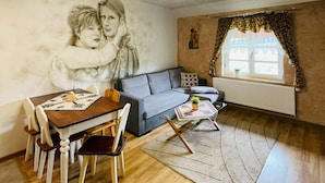 A living room with a sofa, a dining table with chairs, a chandelier, a window with curtains, and a wall with a mural.