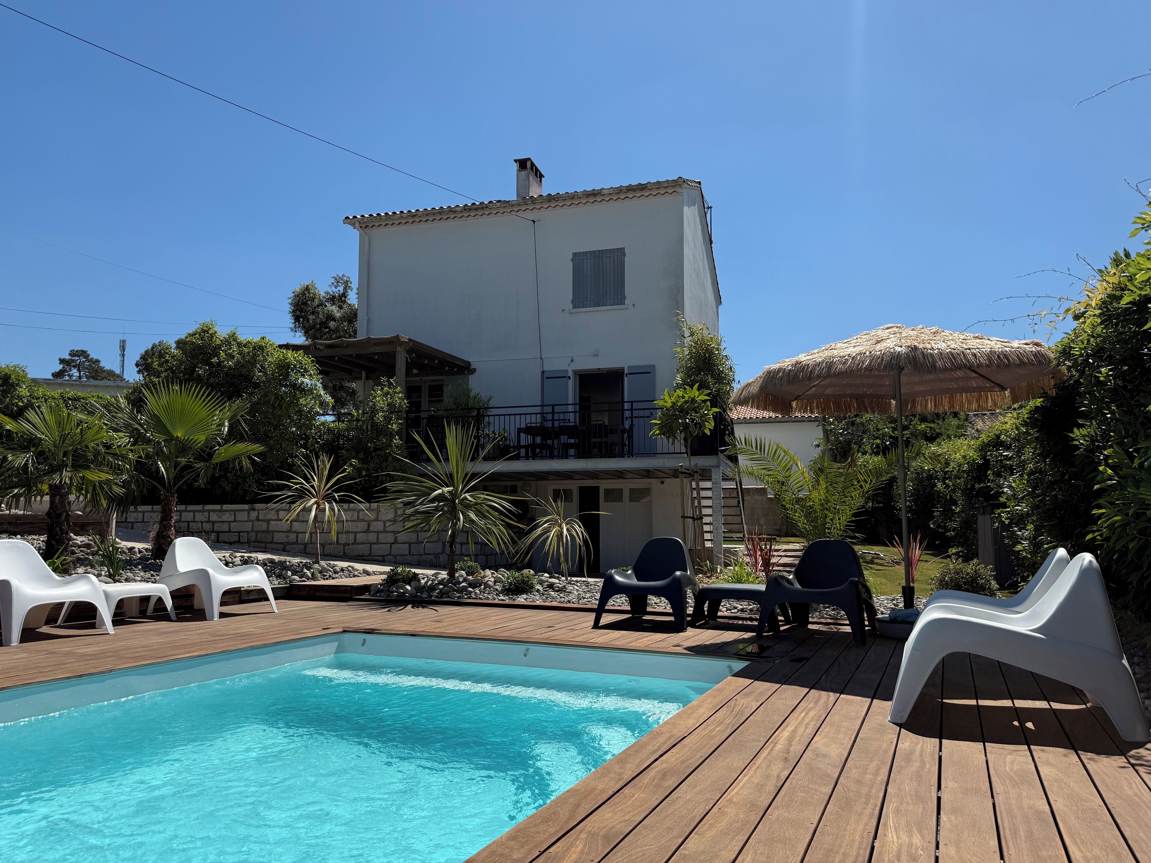 A pool area with a wooden deck, white lounge chairs, and a building with a balcony.
