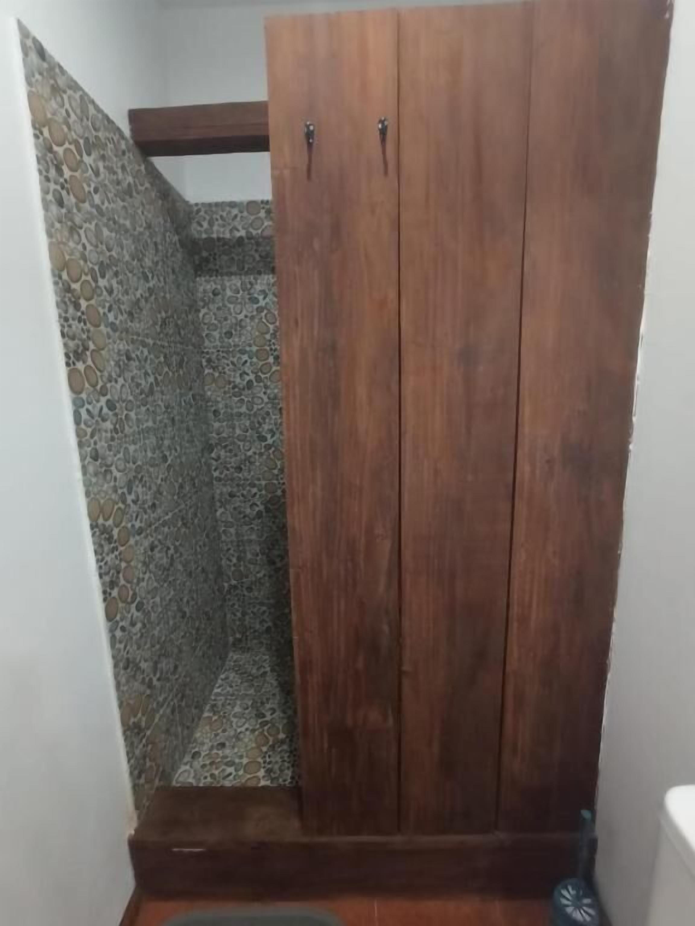 Een houten garderobekast met twee deuren in een hoek van een kamer met een muur met een stenen patroon.