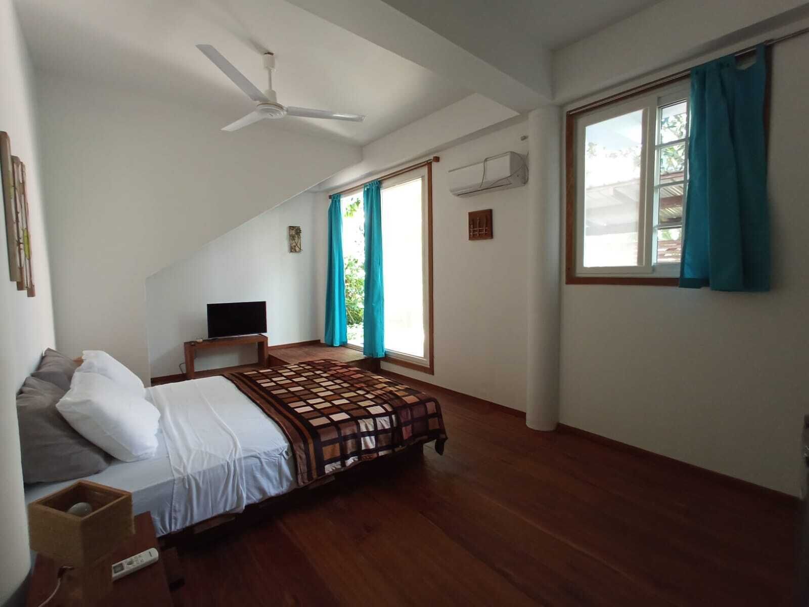 A bedroom with a bed, a TV on a stand, a ceiling fan, and a window with curtains.