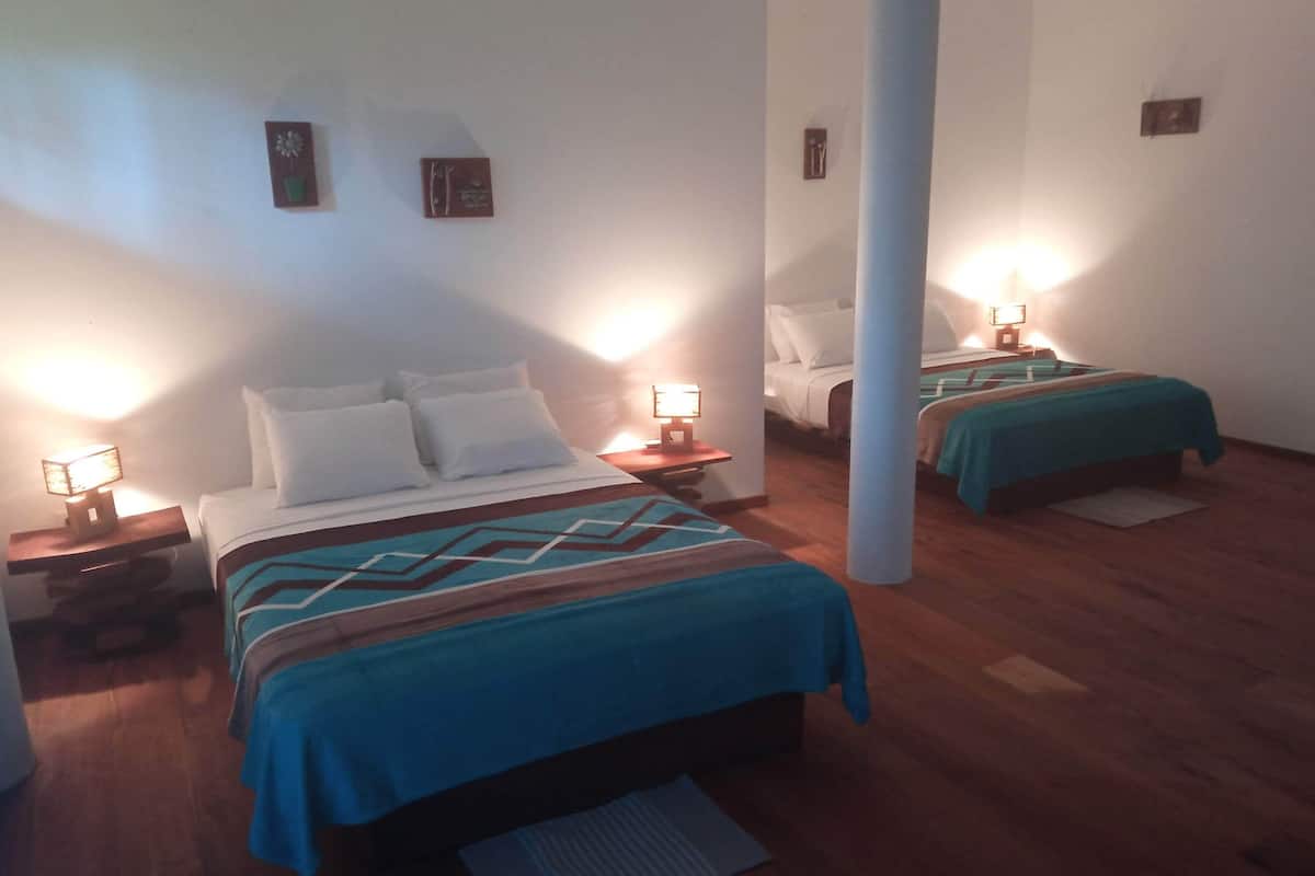 A hotel room with two beds, each with a blue bedspread and white pillows. There are bedside tables with lamps, and the room has wooden flooring.