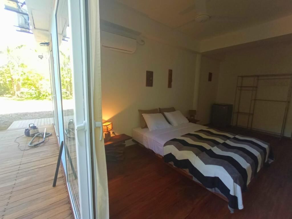 A bedroom with a bed, a wooden floor, a nightstand, a lamp, and a view of the outdoors.