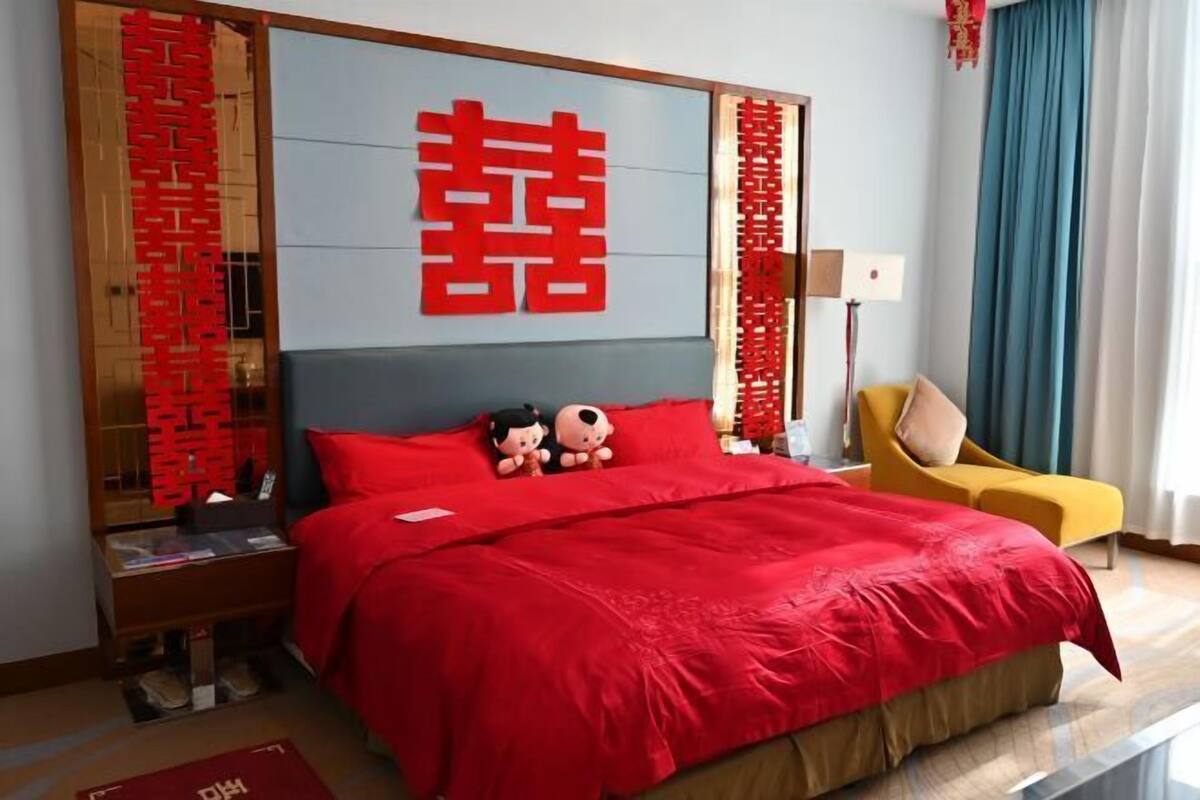Une chambre d’hôtel avec un grand lit, une literie rouge, une table de chevet en bois, un fauteuil jaune et un grand miroir orné de caractères chinois rouges.