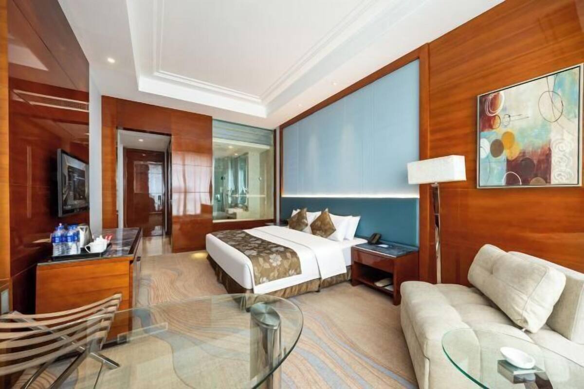Une chambre d’hôtel avec un lit, un canapé, une table en verre, un bureau, une lampe, une télévision et un tableau.