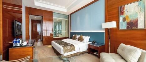 Habitación de hotel con cama, sofá, mesa de vidrio, escritorio, lámpara, televisor y un cuadro.