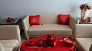Um quarto com um sofá, duas poltronas, uma mesa redonda com toalha vermelha, um bule, xícaras e um vaso com flores.