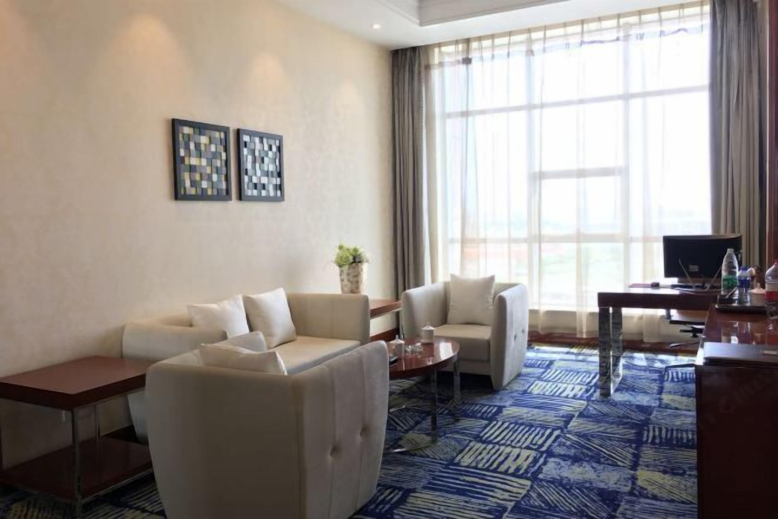 A hotel room with a sofa, armchairs, a coffee table, a desk, a computer, and a large window with curtains.