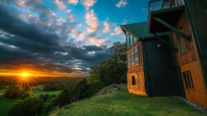 Een houten chalet met een balkon, omgeven door groen en uitzicht op de zonsondergang.