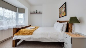 A bedroom with a bed, a nightstand, a lamp, a window with sheer curtains, a radiator, and a painting on the wall.