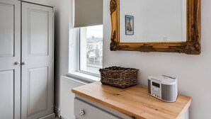 A room with a wooden dresser, a radio, a wicker basket, a mirror with an ornate frame, and a window with a view of buildings.
