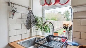 A kitchen with a sink, a dish rack, a hanging pot holder, and a window with a view of a garden.