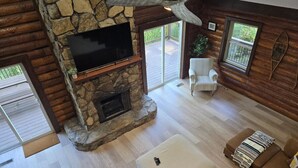 A cozy living room with a stone fireplace, a flat-screen TV, and a comfortable seating area with a cushioned bench and a sofa. - Cozy log cabin, 25 mins to Broadway (Ashland City)