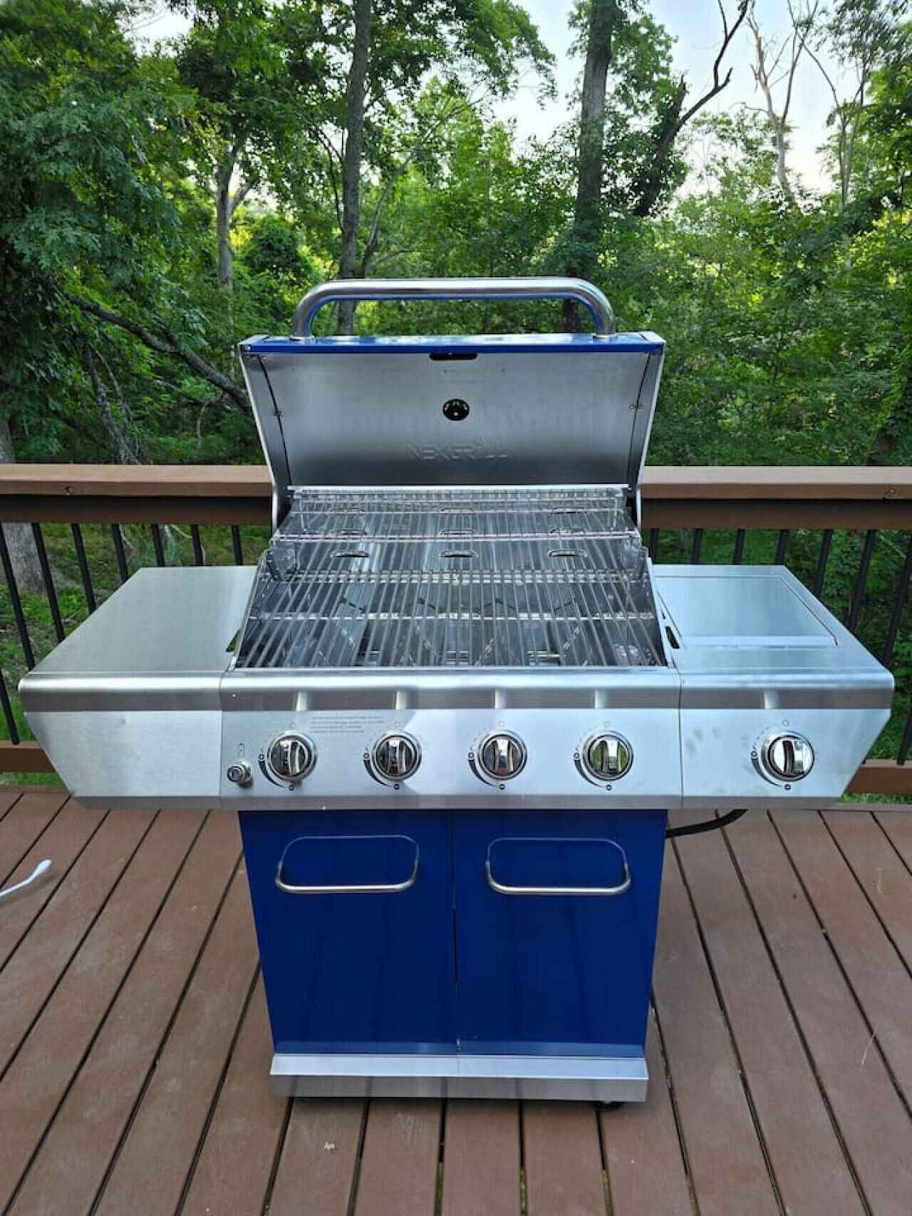 A stainless steel and blue gas grill with four burners, located on a wooden deck with a railing, surrounded by trees.