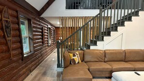 A log cabin interior with a wooden wall, a brown leather sectional sofa, a white ottoman, and a staircase with a wooden railing. - Cozy log cabin, 25 mins to Broadway (Ashland City)