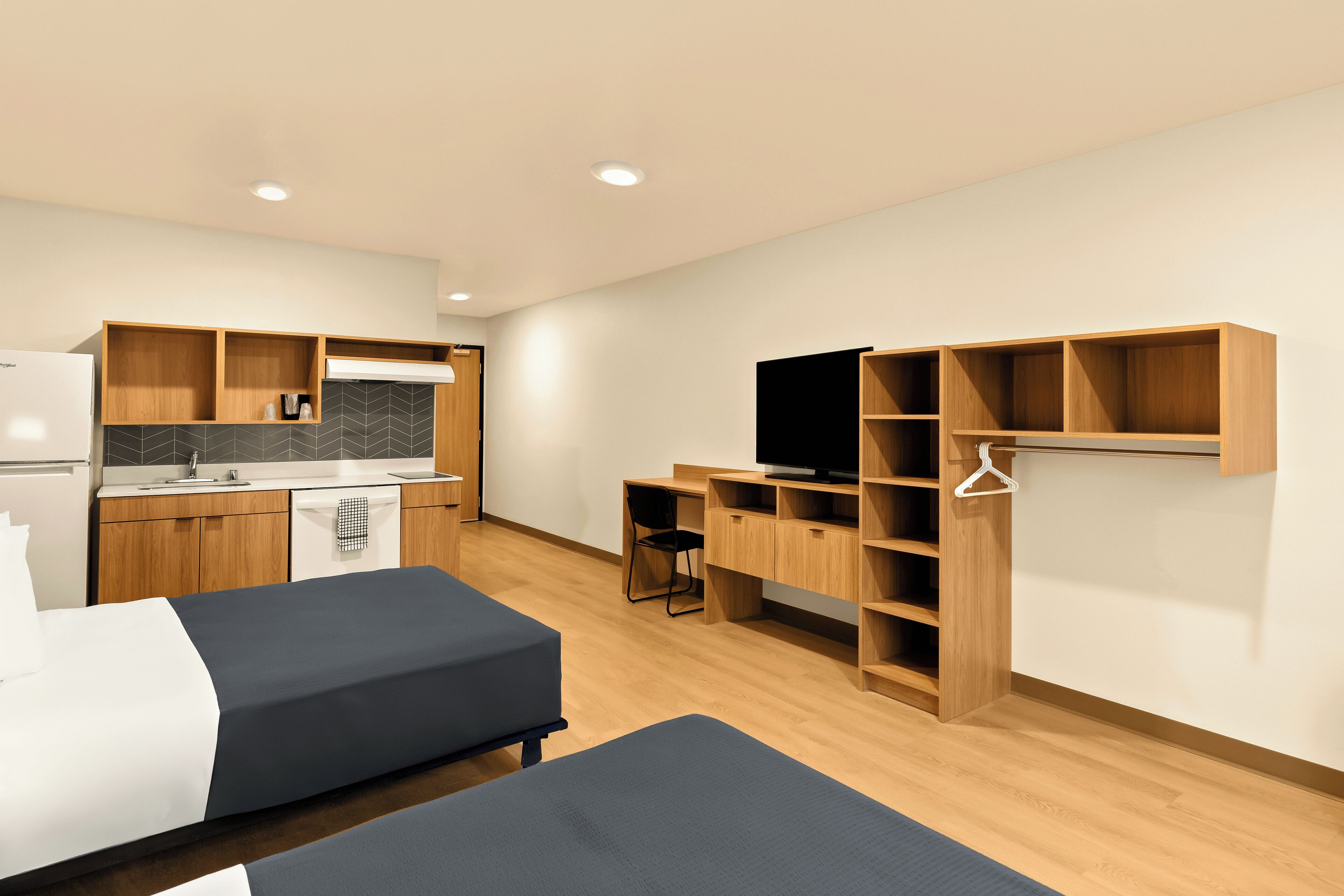 A modern hotel room with a kitchenette, two beds, a desk, and a TV.