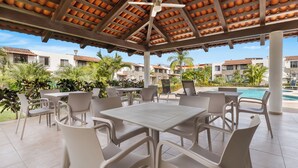 A covered outdoor seating area with white plastic chairs and tables, a ceiling fan, and a view of a swimming pool and buildings in the background. - Modern Pool Side, 3 Bdrm House With Parking (Puerto Vallarta)