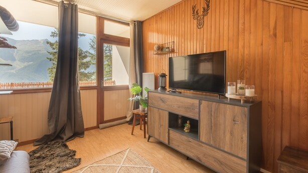 Ein Wohnzimmer mit einer holzverkleideten Wand, einem Flachbildfernseher, einem Kamin und Blick auf Berge durch große Fenster.