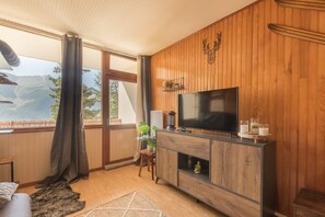 Een woonkamer met een houten wandpaneel, een flatscreen televisie, een open haard en uitzicht op bergen door grote ramen.