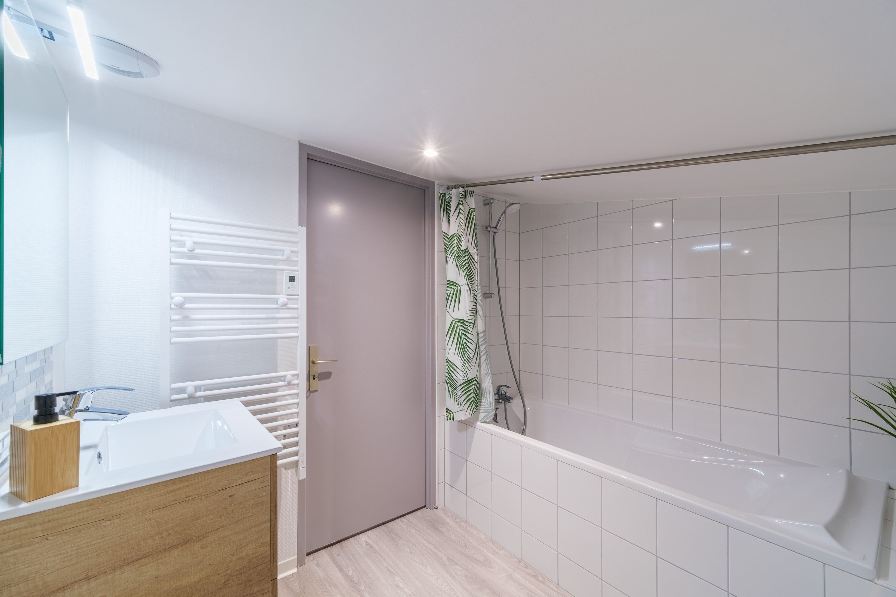 Een moderne badkamer met een witte wastafel, een houten badkamermeubel, een douche met een douchegordijn met bladmotief en een witte badkuip.