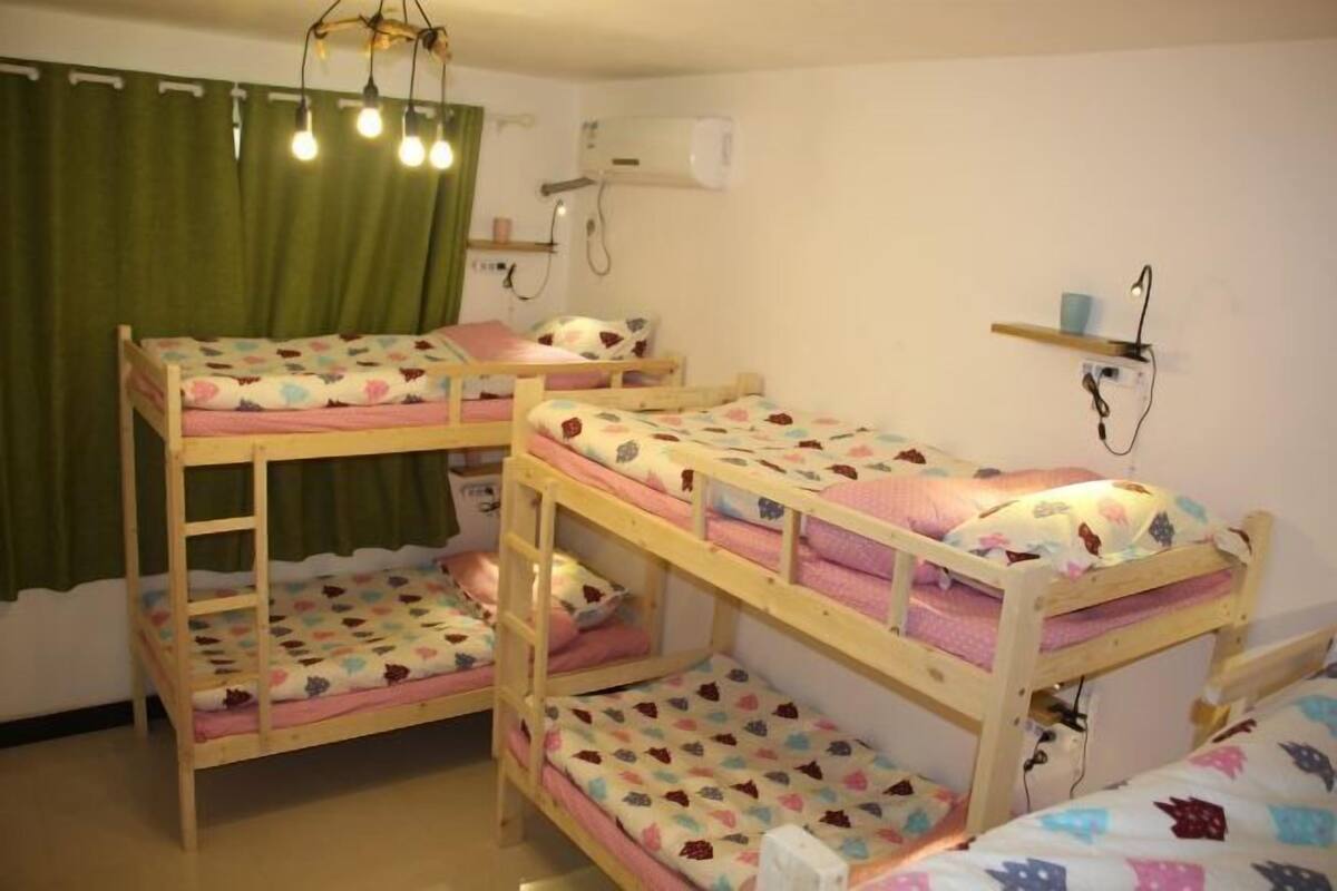 Un dormitorio con camas literas, una cortina verde, un equipo de aire acondicionado y una estantería con una lámpara.