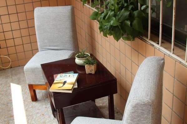 Ein kleines Zimmer mit einem grauen Sessel, einem Holztisch mit einem Buch und einer Pflanze sowie einem Fenster mit einer Pflanze im Fensterbrett.
