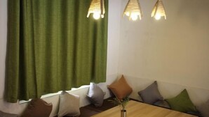A room with a wooden table, a vase with a plant, green curtains, and multiple pillows on a bench.