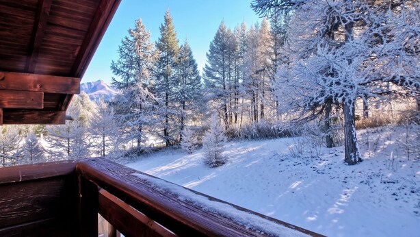 A wooden balcony with a view of a snow-covered forest and mountains.