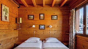 A wooden cabin room with two beds, framed pictures on the wall, a lamp, and a window with curtains. - Casa-Forêt Blanche, splendide chalet (Risoul)
