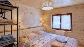 A cozy wooden room with a bed, a shelf with towels, a lamp, and a window.