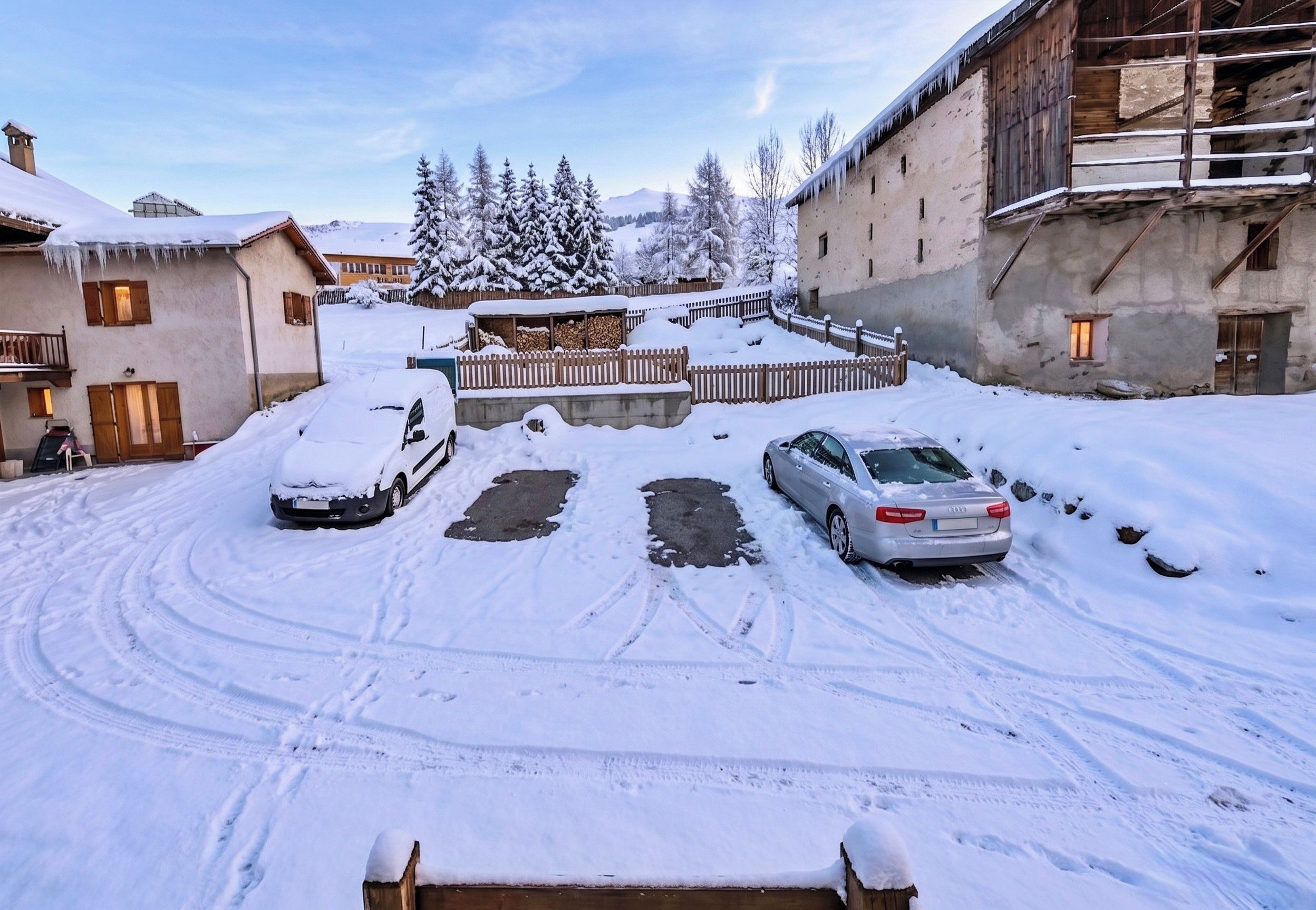 A snow-covered parking area with two cars, a wooden fence, and buildings in the background.