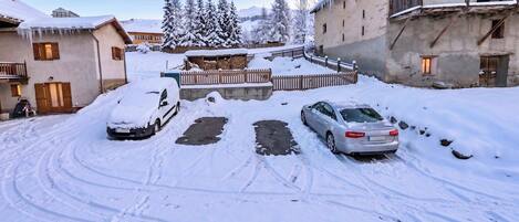 A snow-covered parking area with two cars, a wooden fence, and buildings in the background.
