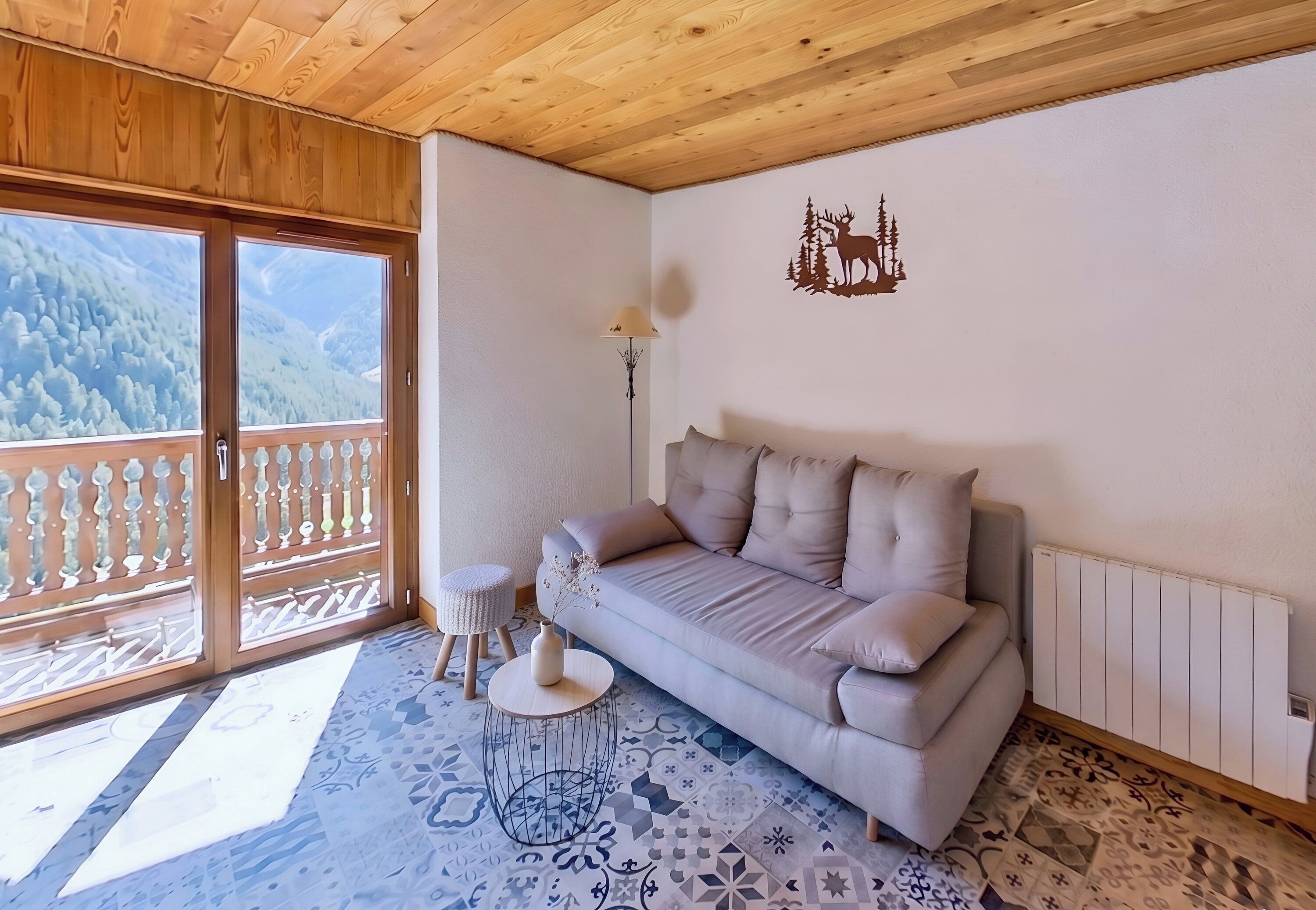 A room with a sofa, a small round table, a lamp, a decorative wall piece, and a view of a forested mountain through a sliding door.