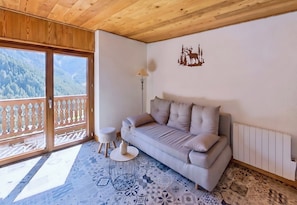 Une pièce comprenant un canapé, une petite table ronde, une lampe, un élément décoratif mural et une vue sur une montagne boisée, visible à travers une porte coulissante.