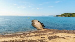 Eine Küstenlandschaft mit einem Betonsteg, der sich in ruhiges blaues Wasser erstreckt, einem Sandstrand und einem fernen, grünen Hügel.
